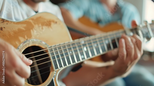 Two guitarists are seen playing their acoustic guitars in a casual setting. One guitar is prominently in the foreground while the other is slightly blurred in the back.