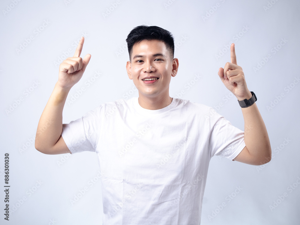 A young Asian man, wearing a white shirt and black watch, looks up while pointing both index fingers upwards. The background is plain white A young Asian man, wearing a white shirt and black watch, looks up while pointing both index fingers upwards. The background is plain white