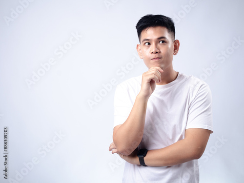 A young Asian man, wearing a white shirt and black watch, looks pensively to the side while resting his chin on his hand. The plain white background emphasizes the subject's contemplative