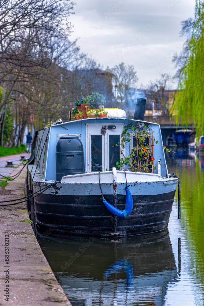 Naklejka premium Beautiful narrow blue wooden boat or houseboat docked and moored in the residential district of Maida Vale in London, with flower pots on its roof and smoke coming from its chimney.
