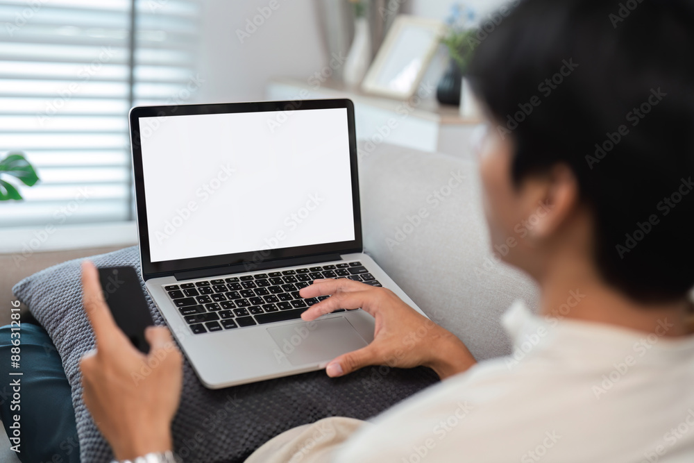 Young Man Relaxing and Working from Home on Laptop with Smartphone in Modern Living Room Young Man Relaxing and Working from Home on Laptop with Smartphone in Modern Living Room
