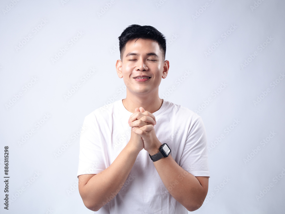 A young Asian man, wearing a white shirt and black watch, smiles peacefully with his hands clasped together in front of him, eyes closed. The background is plain white A young Asian man, wearing a white shirt and black watch, smiles peacefully with his hands clasped together in front of him, eyes closed. The background is plain white