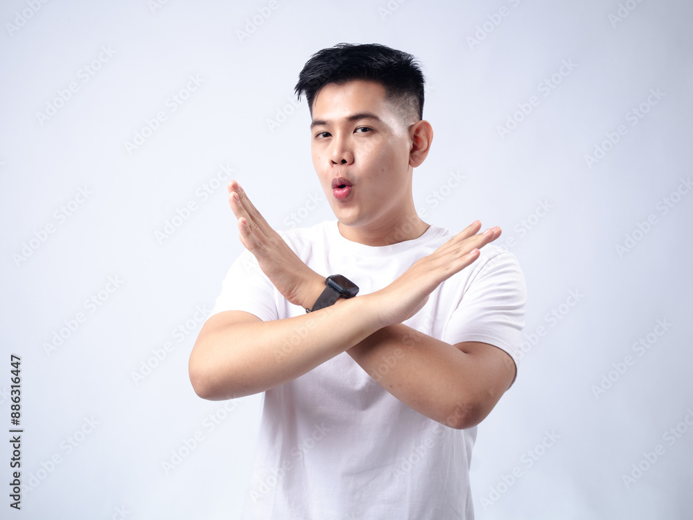 A young Asian man, wearing a white shirt and black watch, crosses his arms in an X shape in front of his chest with a serious expression. The plain white background emphasizes the subject's firm A young Asian man, wearing a white shirt and black watch, crosses his arms in an X shape in front of his chest with a serious expression. The plain white background emphasizes the subject's firm