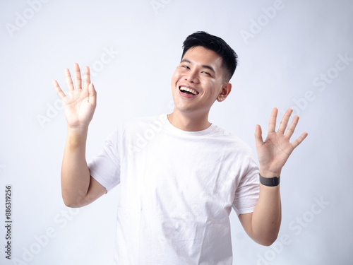 Fotografia A young Asian man, wearing a white shirt and black watch, has a surprised and exaggerated expression with his mouth open and hands spread wide
