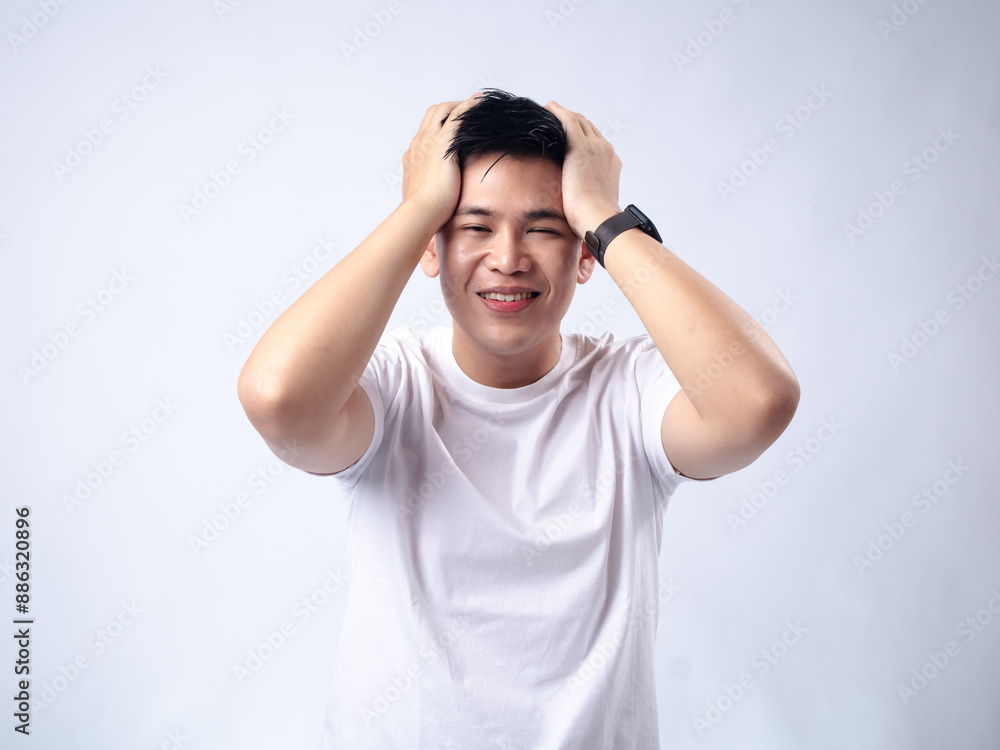 A young Asian man, wearing a white shirt and black watch, looks up thoughtfully while holding his hands behind his head. The background is plain white, highlighting the subject's contemplative A young Asian man, wearing a white shirt and black watch, looks up thoughtfully while holding his hands behind his head. The background is plain white, highlighting the subject's contemplative