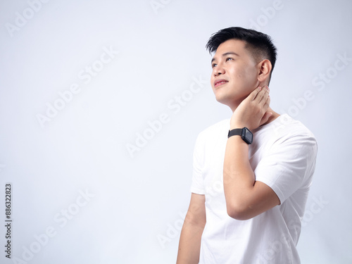 A young Asian man, wearing a white shirt and black watch, looks upwards with a thoughtful expression while touching his neck. The plain white background emphasizes the subject's contemplative