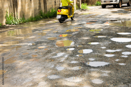 A man rides a yellow scooter on a road with potholes. The road is wet and the potholes are filled with water
