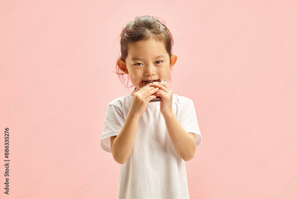 Photo of Cute Little Asian Girl Standing on a Pink Isolated Background, Eating a Chocolate Bar With an Appetite. Sweet Tooth Dilemma. Kids' Dental Health Alert. Photo of Cute Little Asian Girl Standing on a Pink Isolated Background, Eating a Chocolate Bar With an Appetite. Sweet Tooth Dilemma. Kids' Dental Health Alert.