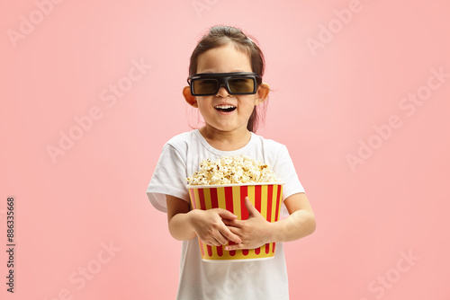 Joyful and Surprised Child Girl in 3D Glasses Holding Popcorn Bucket Standing Over Pink Isolated Background. photo