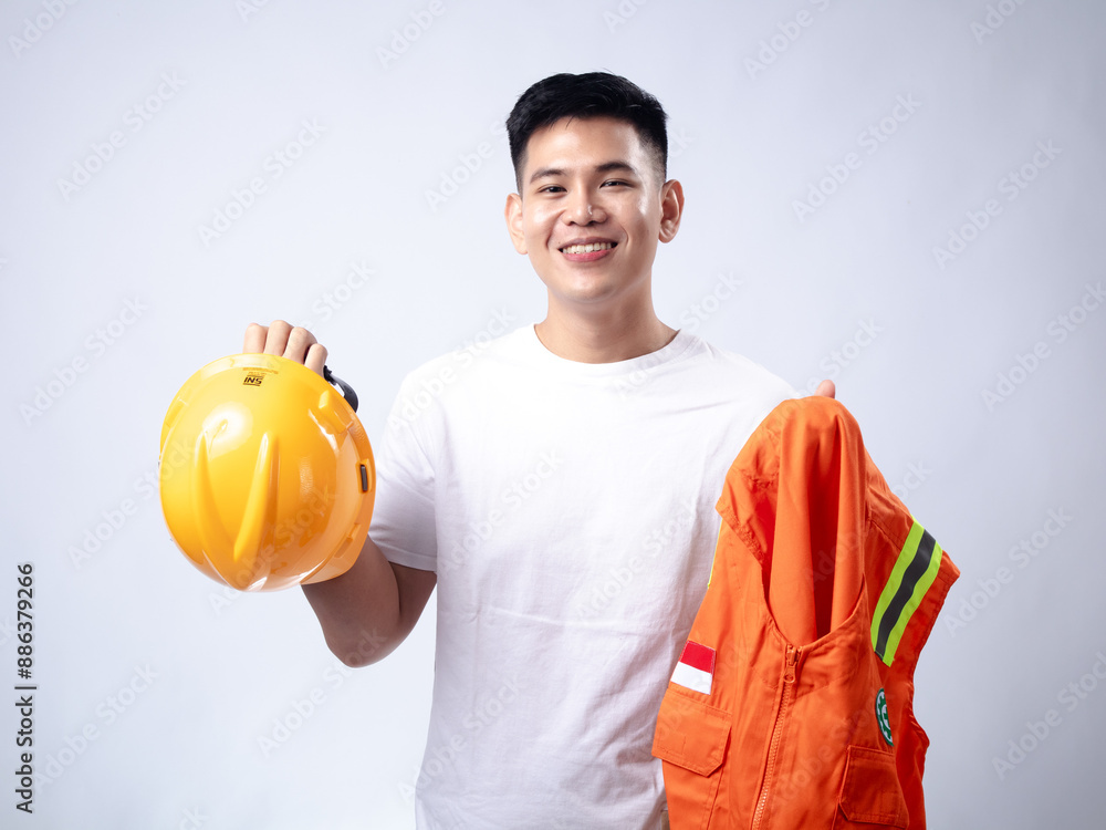 Young male construction worker in a white t-shirt, smiling while holding a yellow hard hat and an orange high-visibility vest. The white background accentuates his cheerful expression for work Young male construction worker in a white t-shirt, smiling while holding a yellow hard hat and an orange high-visibility vest. The white background accentuates his cheerful expression for work