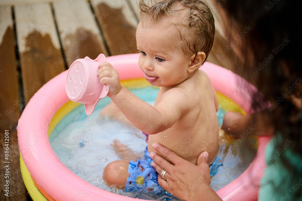 Baby playing with watering can in inflatable pool under adult supervision Baby playing with watering can in inflatable pool under adult supervision