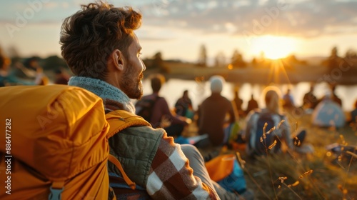 A young individual with a bright orange backpack sits by a lakeside, surrounded by a group, all embracing the serene sunset. The golden hour enhances the peaceful ambiance. photo