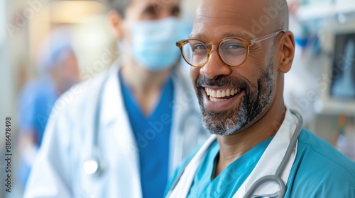 A male doctor, with glasses and a stethoscope around his neck, smiles warmly as his colleagues work in the background of a busy hospital environment.