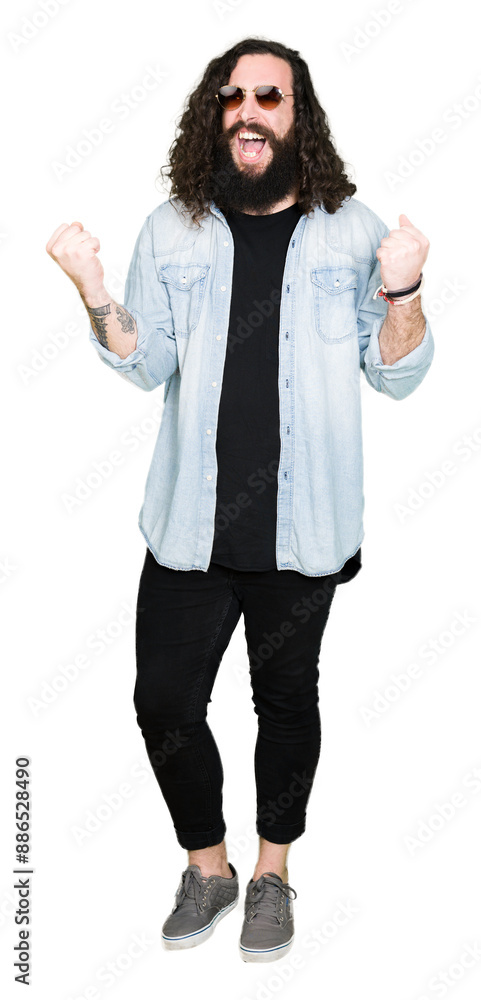 Young hipster man with long hair and beard wearing sunglasses very happy and excited doing winner gesture with arms raised, smiling and screaming for success. Celebration concept.