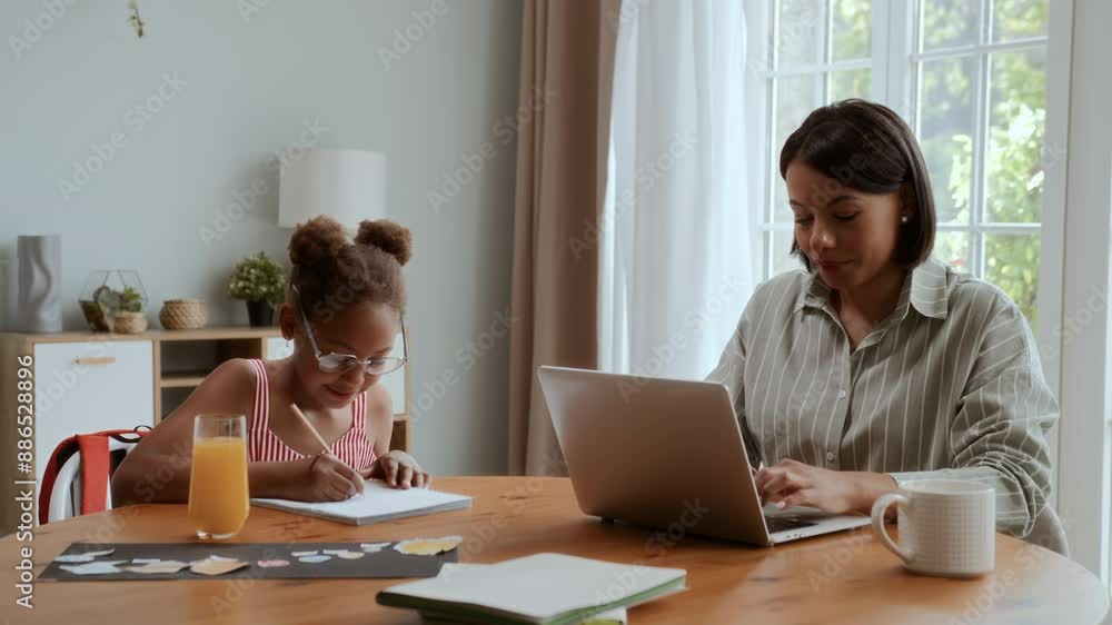 African American mother and daughter sitting at table at home. Black girl is drawing and puts on moms eyeglasses, woman is working on laptop and talks with kid smiling and enjoying African American mother and daughter sitting at table at home. Black girl is drawing and puts on moms eyeglasses, woman is working on laptop and talks with kid smiling and enjoying