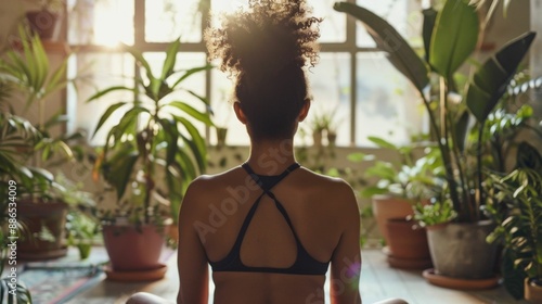 A person with curly hair wearing a black strappy top sitting cross-legged on a wooden floor surrounded by potted plants in a room with natural light. photo