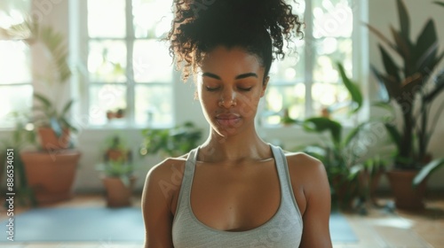 A woman in a tank top with her eyes closed standing in a room with natural light and potted plants appearing to be in a meditative or peaceful state. Free Download A woman in a tank top with her eyes closed standing in a room with natural light and potted plants appearing to be in a meditative or peaceful state. - GFXLoad