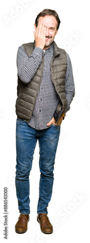 Middle age handsome man wearing winter vest covering one eye with hand with confident smile on face and surprise emotion.