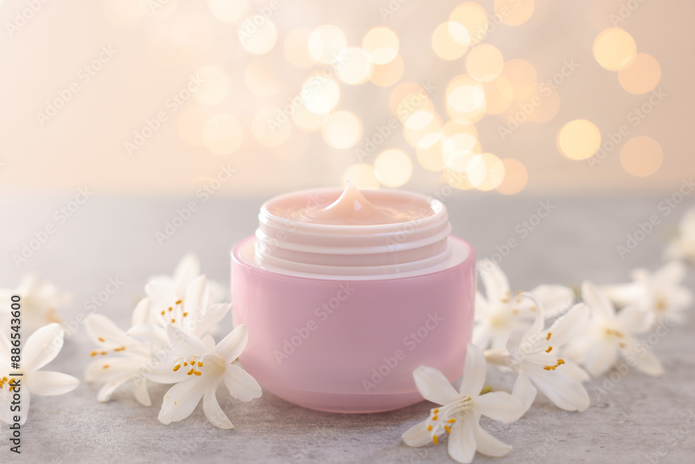 Cream in jar and beautiful jasmine flowers on grey table against light background with blurred lights, closeup