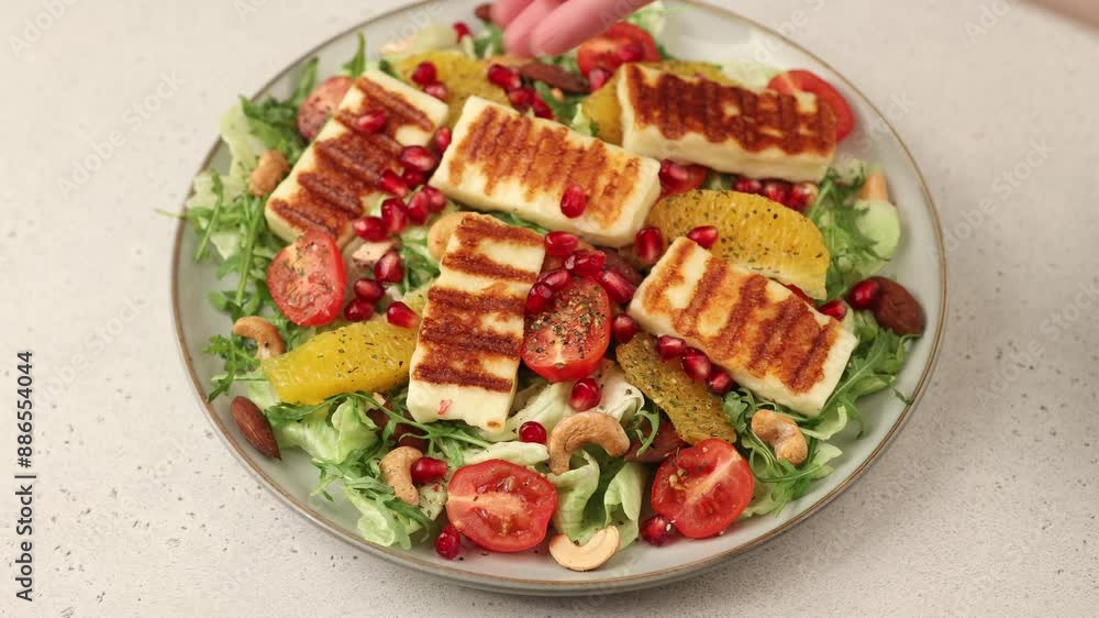 Warm salad with fried halloumi cheese, nuts, cherry tomatoes, pomegranate, orange and arugula. A woman sprinkles pomegranate seeds on a salad. Traditional cheese of the Eastern Mediterranean. Warm salad with fried halloumi cheese, nuts, cherry tomatoes, pomegranate, orange and arugula. A woman sprinkles pomegranate seeds on a salad. Traditional cheese of the Eastern Mediterranean.