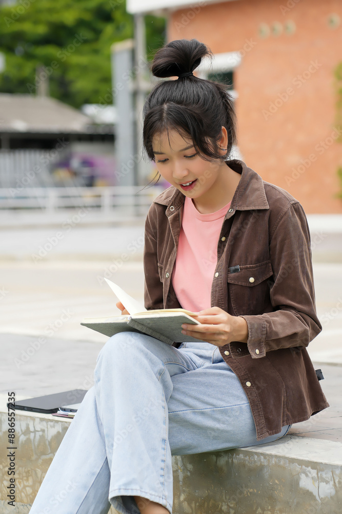 Fototapeta premium A high school student walks outside a school building, looking at her phone. She is smiling and appears engaged with her phone