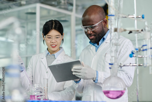Fotografie Two young intercultural chemists looking at screen of tablet held by African Ame