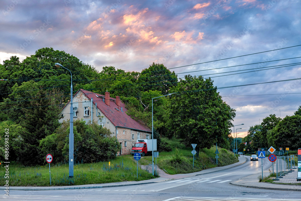 A house with a red tiled roof standing among the trees of a city park against the backdrop of a beautiful sunset sky. Crossroads with road signs. A house with a red tiled roof standing among the trees of a city park against the backdrop of a beautiful sunset sky. Crossroads with road signs.