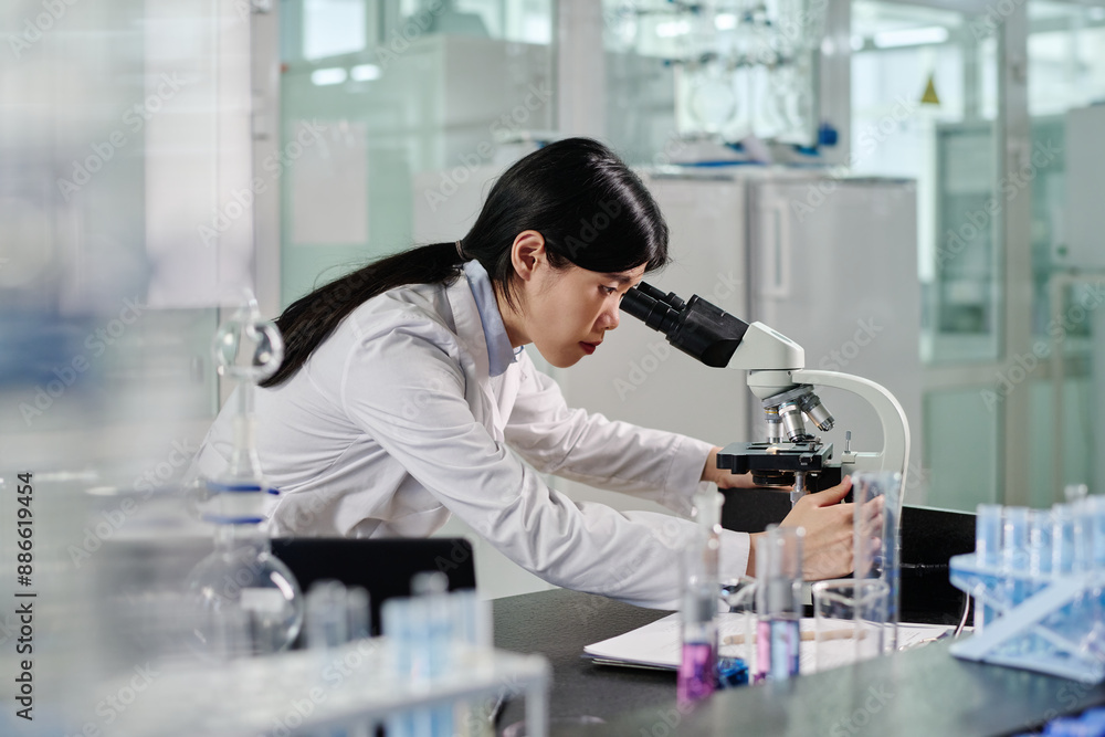 Side view of young Chinese woman in lab coat bending over workplace with variety of glassware and looking in microscope Side view of young Chinese woman in lab coat bending over workplace with variety of glassware and looking in microscope