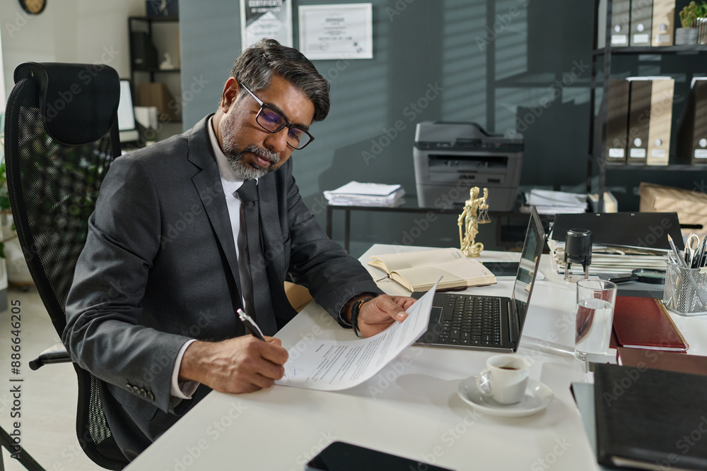 Serious mature lawyer in formalwear sitting by desk with laptop, cup of tea and office supplies and putting his signature in contract Serious mature lawyer in formalwear sitting by desk with laptop, cup of tea and office supplies and putting his signature in contract
