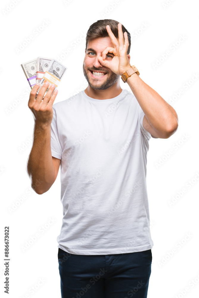 Young handsome man holding stack of dollars over isolated background with happy face smiling doing ok sign with hand on eye looking through fingers Young handsome man holding stack of dollars over isolated background with happy face smiling doing ok sign with hand on eye looking through fingers