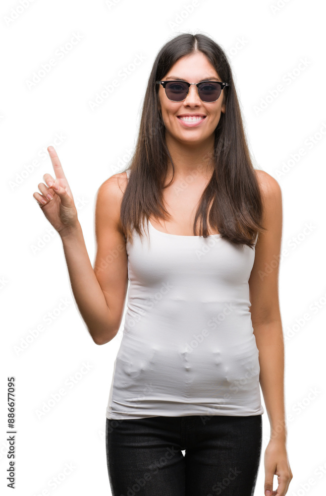Young beautiful hispanic wearing sunglasses showing and pointing up with finger number one while smiling confident and happy. Young beautiful hispanic wearing sunglasses showing and pointing up with finger number one while smiling confident and happy.