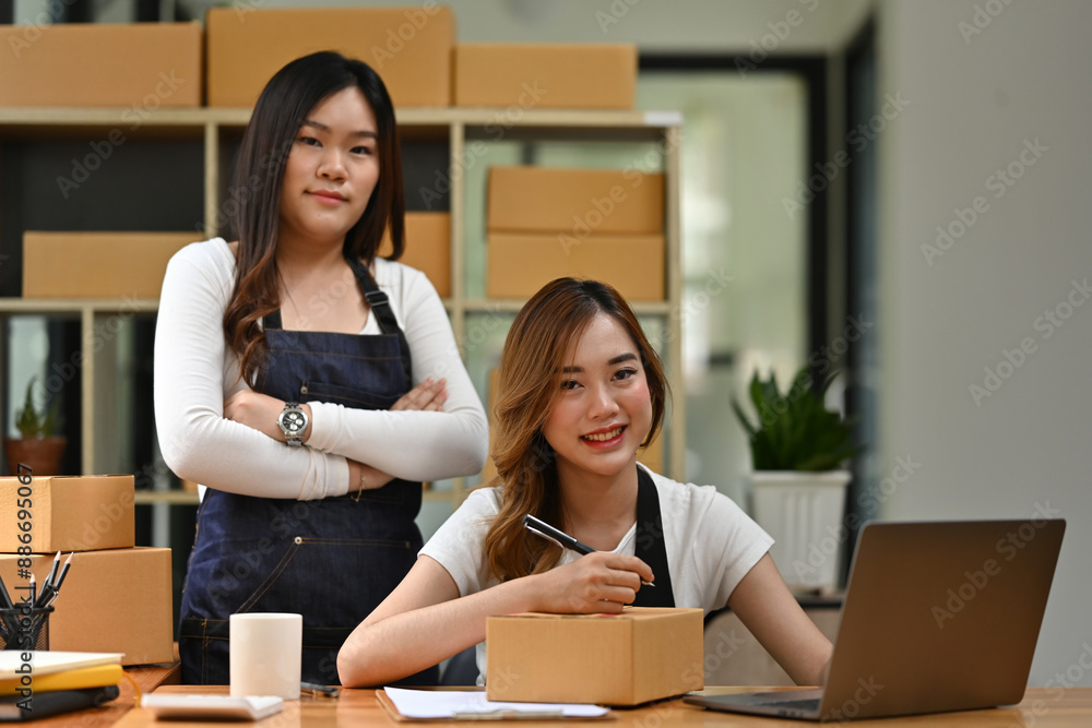 Small Business Teamwork, Two women confidently manage their small business, with one labeling a package and the other standing proudly beside, the SME concept Small Business Teamwork, Two women confidently manage their small business, with one labeling a package and the other standing proudly beside, the SME concept