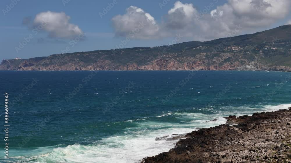 A view of the Atlantic Ocean crashing against the rocky coastline of Guincho, Portugal. Whitecaps and foamy waves break against the rugged rocks, while a distant coastline is visible in the background A view of the Atlantic Ocean crashing against the rocky coastline of Guincho, Portugal. Whitecaps and foamy waves break against the rugged rocks, while a distant coastline is visible in the background