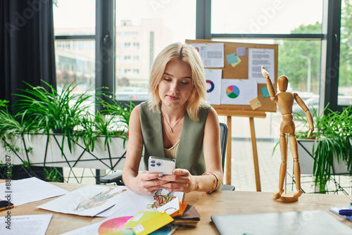Εκτύπωση καμβά A blonde woman in a green sleeveless shirt is sitting at a desk in an office, checking her phone and looking at designs on the table