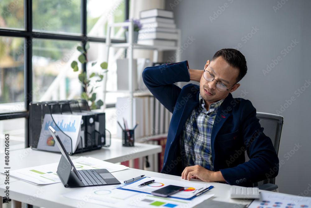 A man in a blue jacket is hunched over a desk with a laptop and a cell phone A man in a blue jacket is hunched over a desk with a laptop and a cell phone