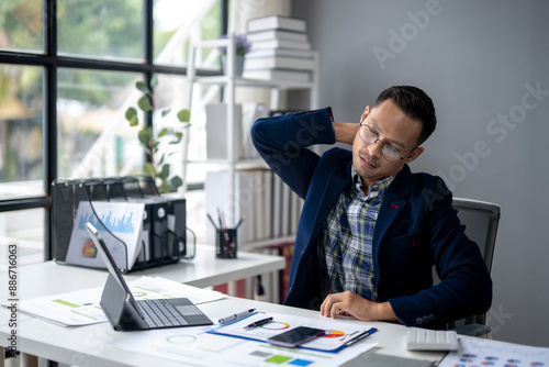 A man in a blue jacket is hunched over a desk with a laptop and a cell phone