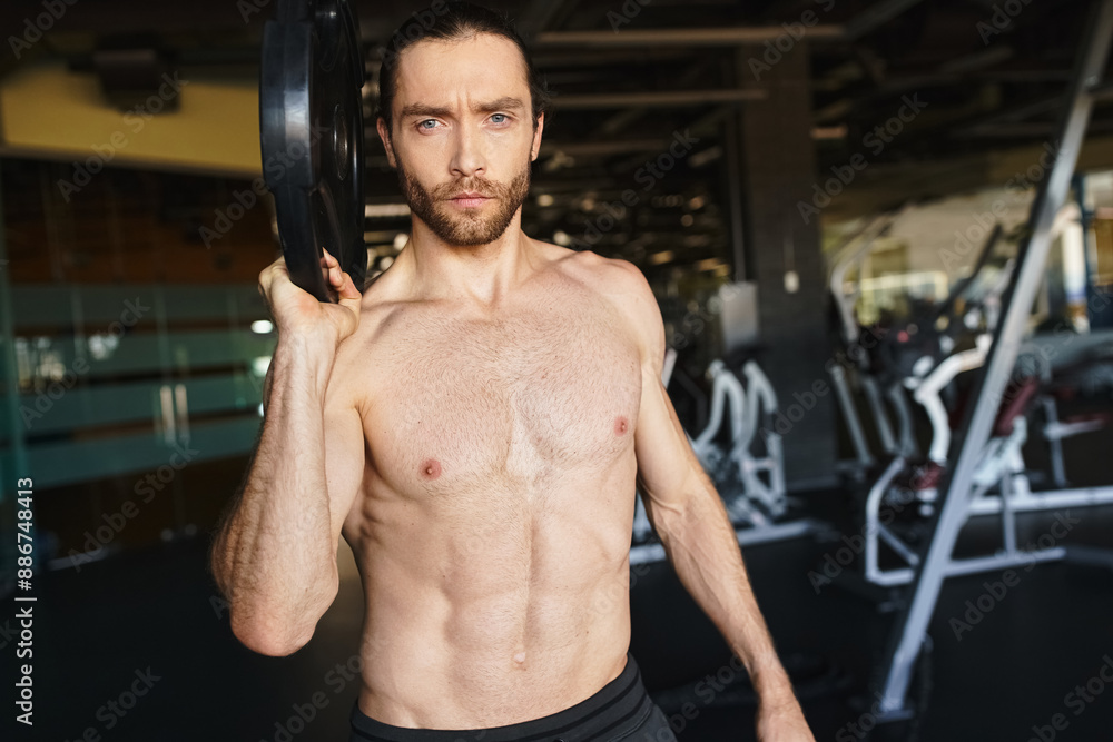 Shirtless man showcasing strength, holding a black weight plate in gym. Shirtless man showcasing strength, holding a black weight plate in gym.