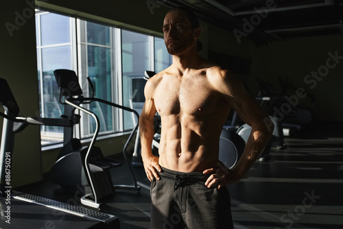 A muscular man without a shirt standing in front of a gym machine, focused and ready to workout.