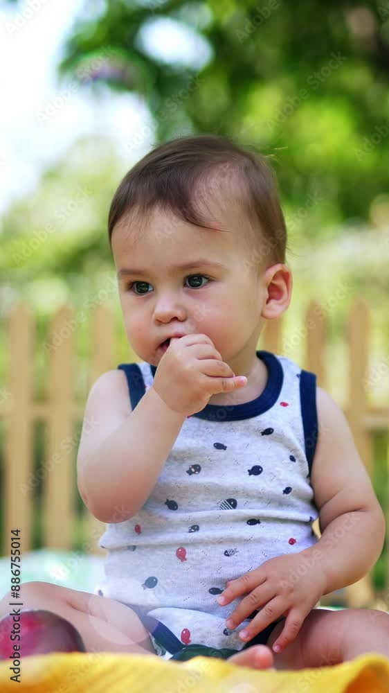 Lovely child in light summer clothes sitting in the garden. Kid holding thumb in mouth looking ahead not noticing flying soap bubbles. Vertical video Lovely child in light summer clothes sitting in the garden. Kid holding thumb in mouth looking ahead not noticing flying soap bubbles. Vertical video
