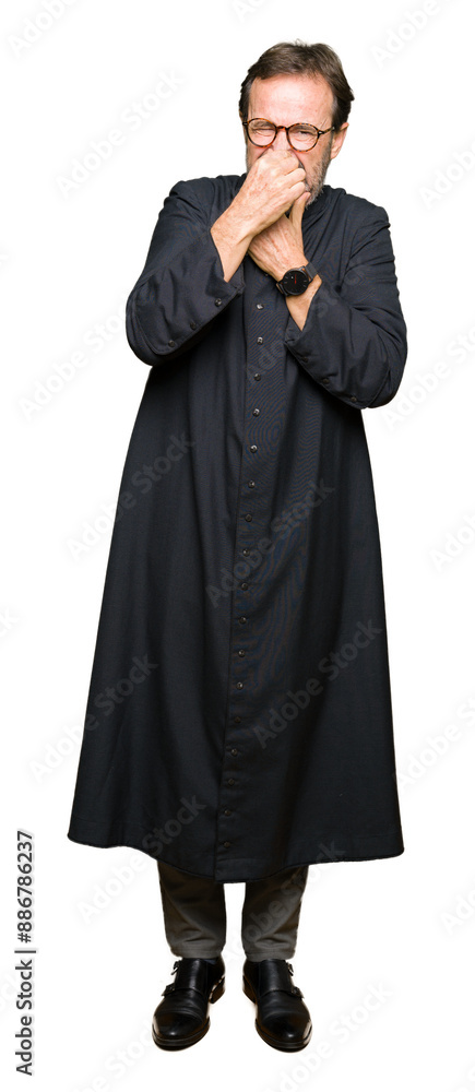 Middle age priest man wearing catholic robe smelling something stinky and disgusting, intolerable smell, holding breath with fingers on nose. Bad smells concept. Middle age priest man wearing catholic robe smelling something stinky and disgusting, intolerable smell, holding breath with fingers on nose. Bad smells concept.