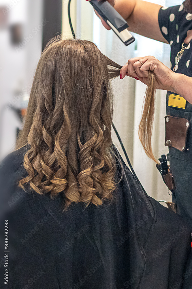 Naklejka premium Hair stylist prepares woman makes curls hairstyle with curling iron. Long light brown natural hair