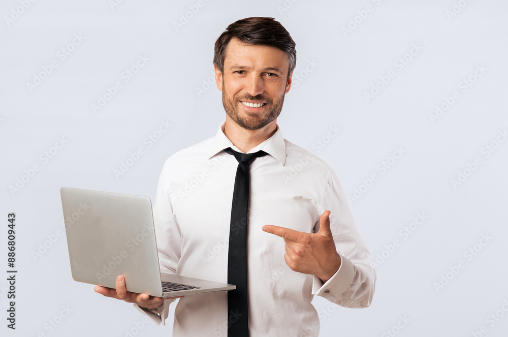 A man in a white shirt and black tie is holding a laptop in his left hand and pointing to the right with his right hand. He is smiling and looking directly at the camera. A man in a white shirt and black tie is holding a laptop in his left hand and pointing to the right with his right hand. He is smiling and looking directly at the camera.