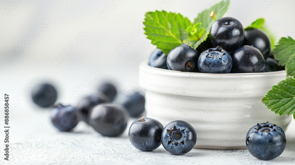 A white bowl holds blueberries, surrounded by green leaves on a white table A white bowl holds blueberries, surrounded by green leaves on a white table