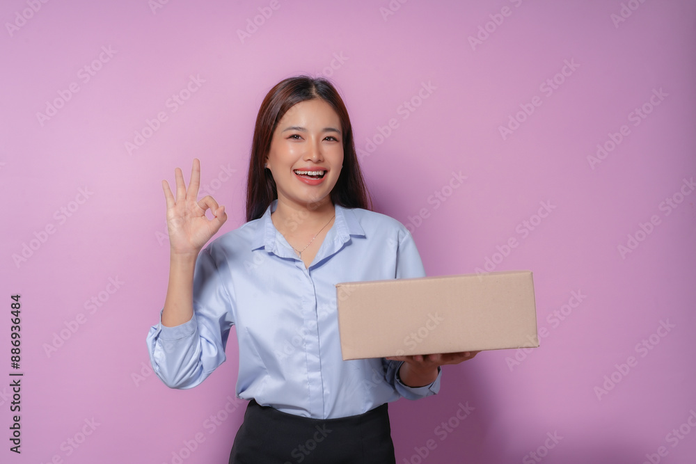Young woman in a studio setting, holding a cardboard box and flashing the ok sign, radiates happiness with a cheerful smile Young woman in a studio setting, holding a cardboard box and flashing the ok sign, radiates happiness with a cheerful smile
