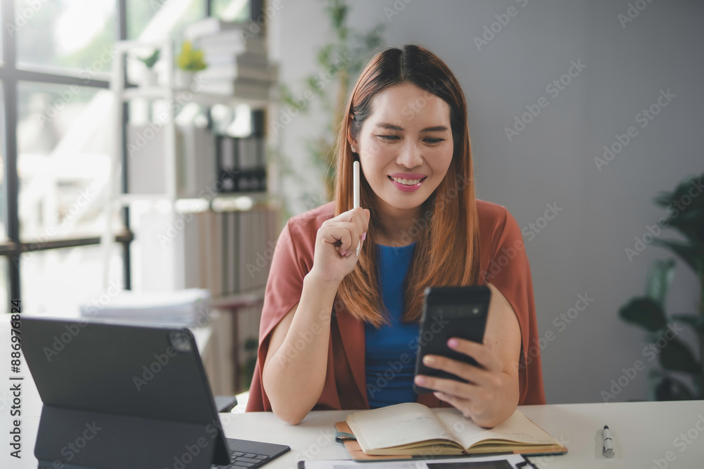 Young businesswoman at her desk, using her smartphone and holding a pen while deep in thought, possibly planning a new strategy or creative idea in a modern office setting Young businesswoman at her desk, using her smartphone and holding a pen while deep in thought, possibly planning a new strategy or creative idea in a modern office setting
