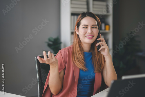 Asian businesswoman multitasks in her office, talking on the phone and gesturing while working on her laptop, showcasing her confidence and determination in communication and technology photo