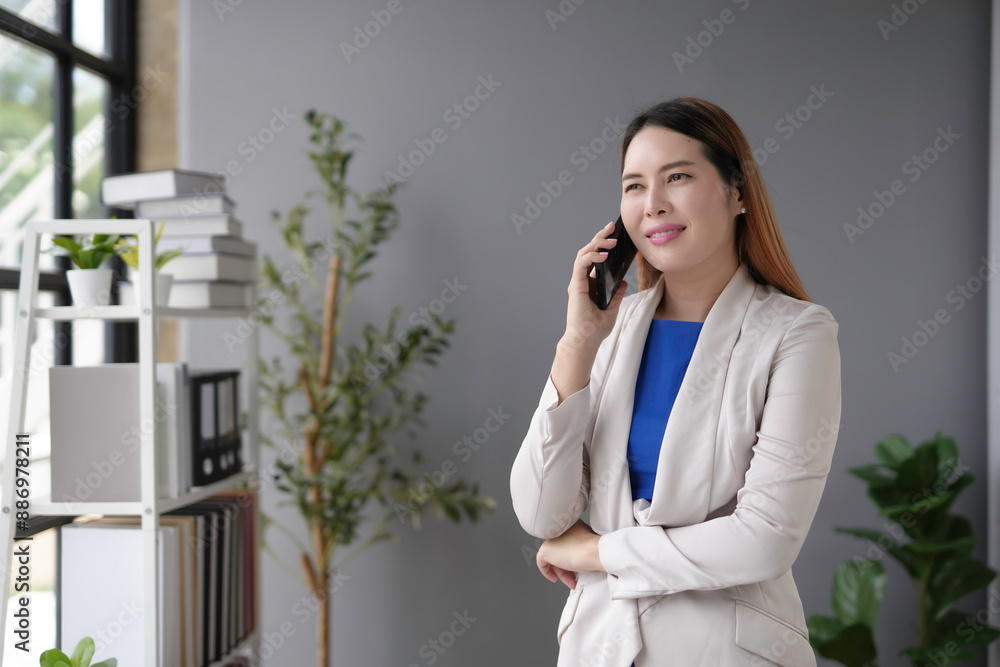 Confident asian businesswoman in modern office, smiling on phone call. Bright blue interior with window, plant, bookshelf. Success and professionalism shown with mobile device Confident asian businesswoman in modern office, smiling on phone call. Bright blue interior with window, plant, bookshelf. Success and professionalism shown with mobile device