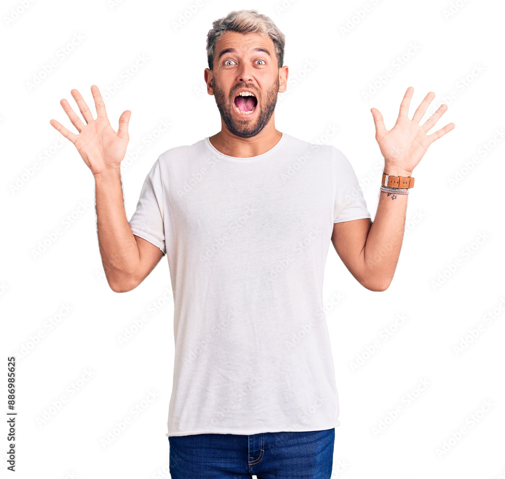 Young handsome blond man wearing casual t-shirt celebrating surprised and amazed for success with arms raised and open eyes. winner concept. Young handsome blond man wearing casual t-shirt celebrating surprised and amazed for success with arms raised and open eyes. winner concept.