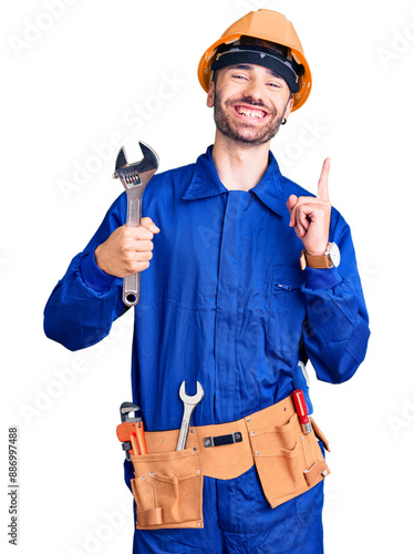 Young hispanic man wearing electrician uniform holding wrench surprised with an idea or question pointing finger with happy face, number one photo