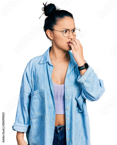 Beautiful hispanic woman wearing casual denim jacket and glasses looking stressed and nervous with hands on mouth biting nails. anxiety problem.